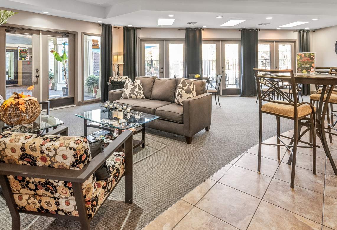 A cozy and well-lit living room, furnished with a couch, coffee table, and chair, has a large window allowing natural light to brighten the space, and a potted plant near the couch providing a touch of greenery.